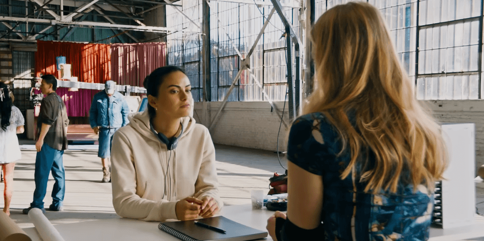 Two women talking in a sunlit warehouse with people and equipment in the background.