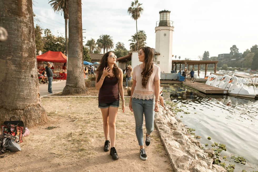 Two women walking by a lakeside lighthouse, with boats and market stalls nearby.