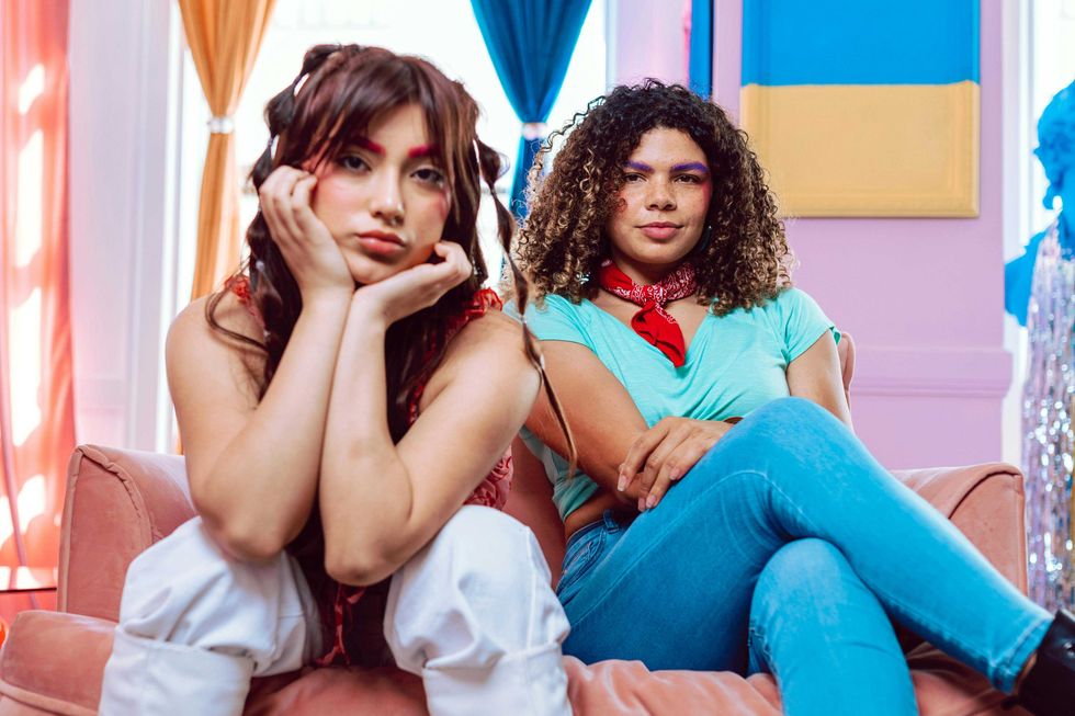 Two women with colorful makeup sitting on a pink couch in a vibrant, colorful room.