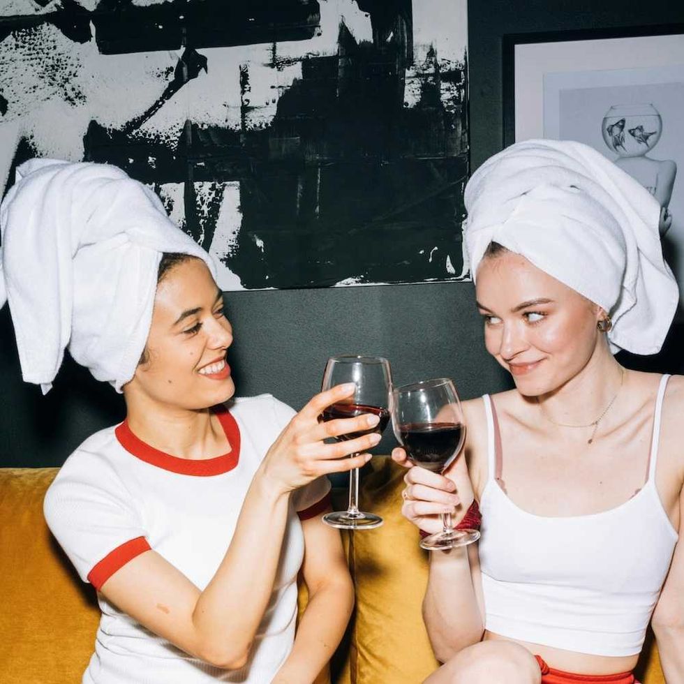 Two women with towel turbans toast with red wine on a couch.