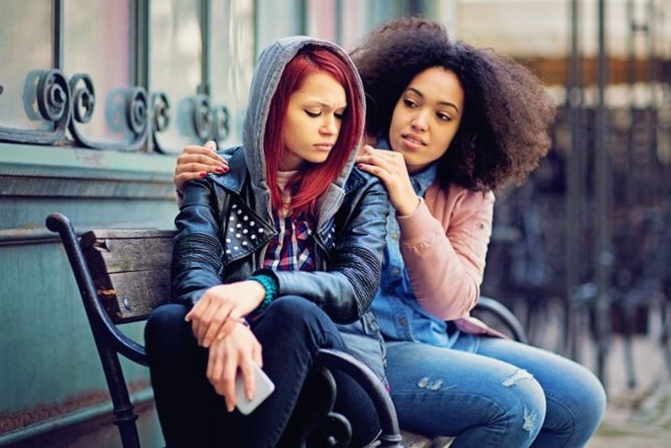 Two young women sit on a bench as one consoles the other
