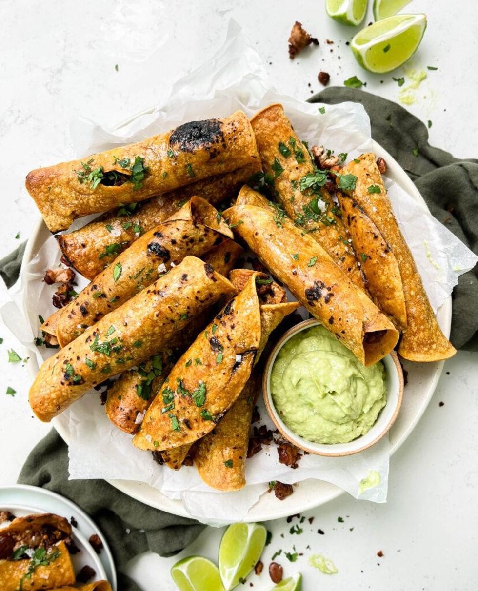 Vegan taquitos are piled on a plate with guac.