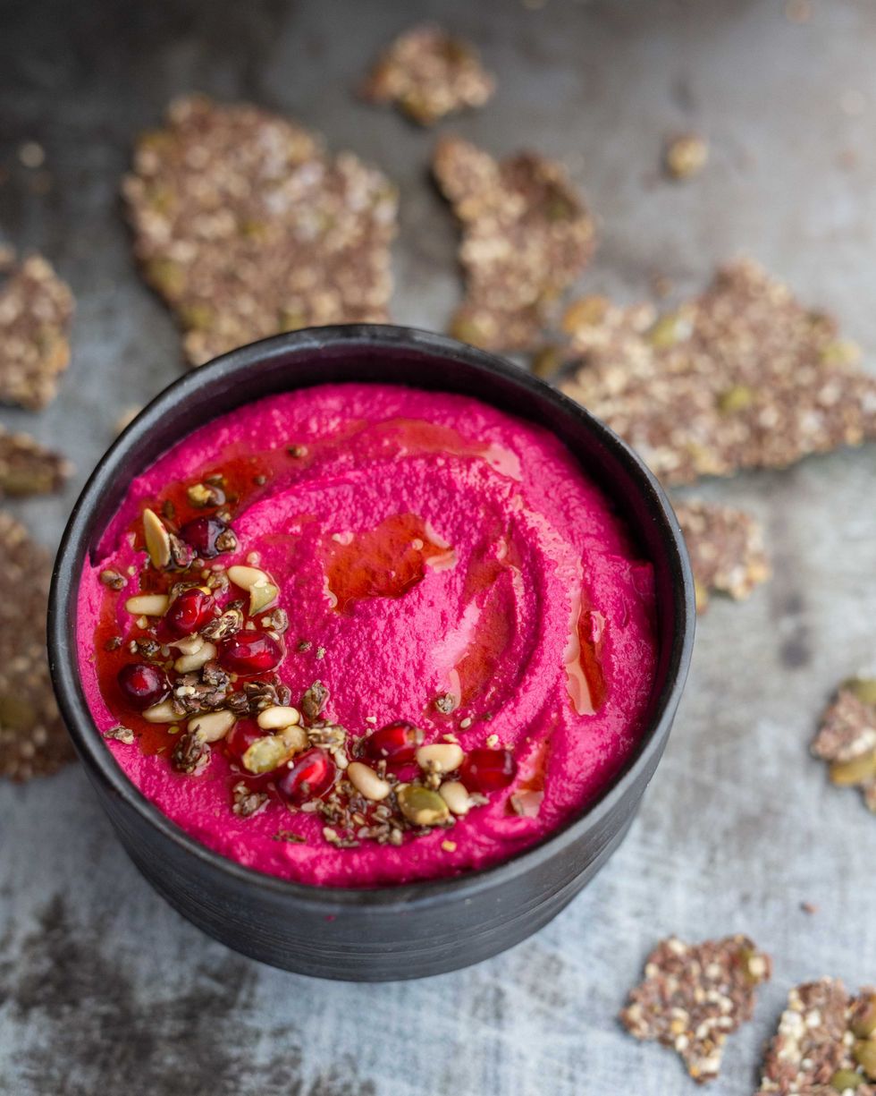 Vibrant beet hummus in a black bowl, garnished with seeds and pomegranate arils.