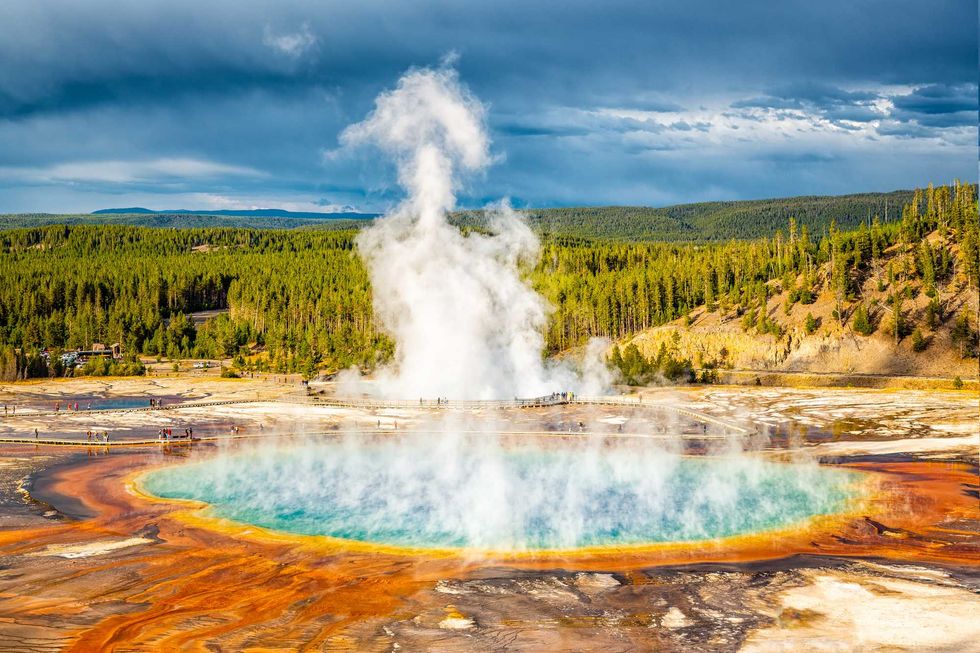 Vibrant hot spring with steam, surrounded by forest under a dramatic sky.