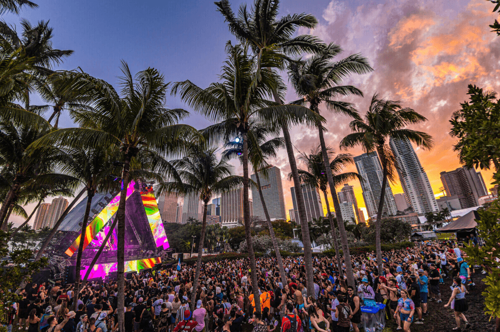 Vibrant music festival with a crowd, palm trees, and city skyline at sunset.