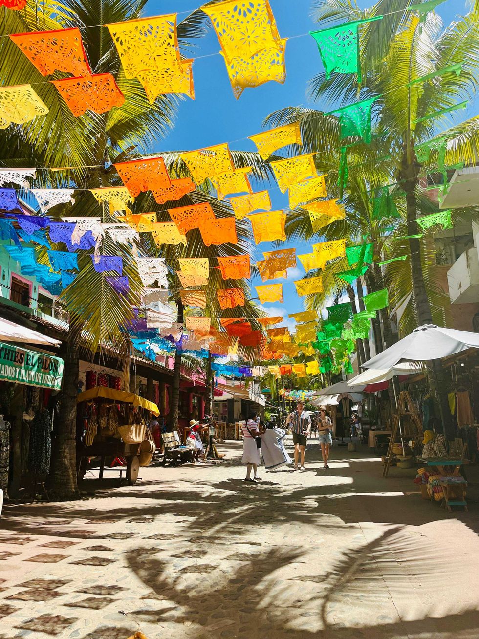 Vibrant papel picado hang over a sunny, bustling street with palm trees and market stalls.