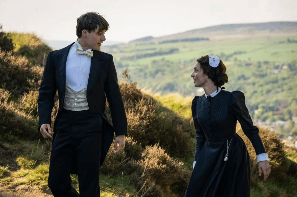 Victorian-era couple walking on a grassy hill in formal attire, sharing a smile.