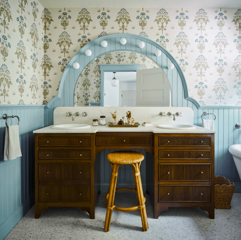 Vintage bathroom with floral wallpaper, wooden vanity, and round mirror.