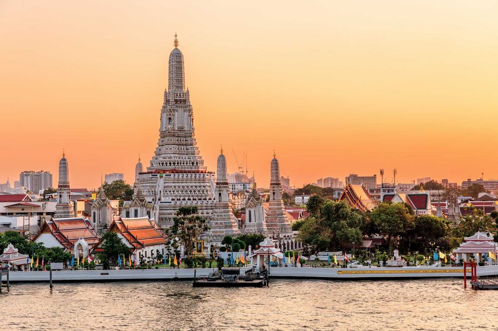 Wat Arun temple at sunset with orange sky, Bangkok skyline, and Chao Phraya River in view.