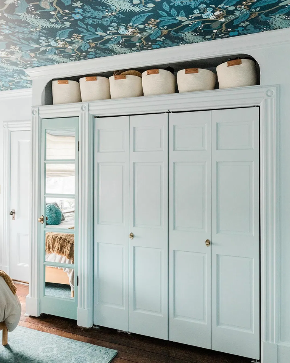White closet with baskets on top, mirrored door, and floral ceiling wallpaper.