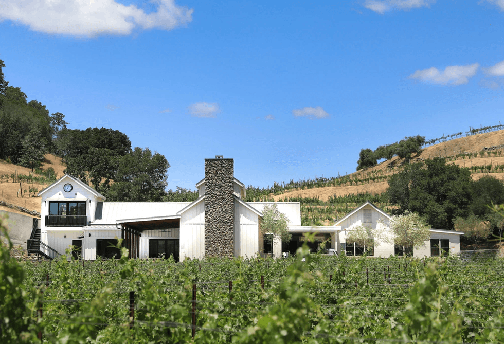 White farmhouse amidst vineyards, with a blue sky and rolling hills in the background.