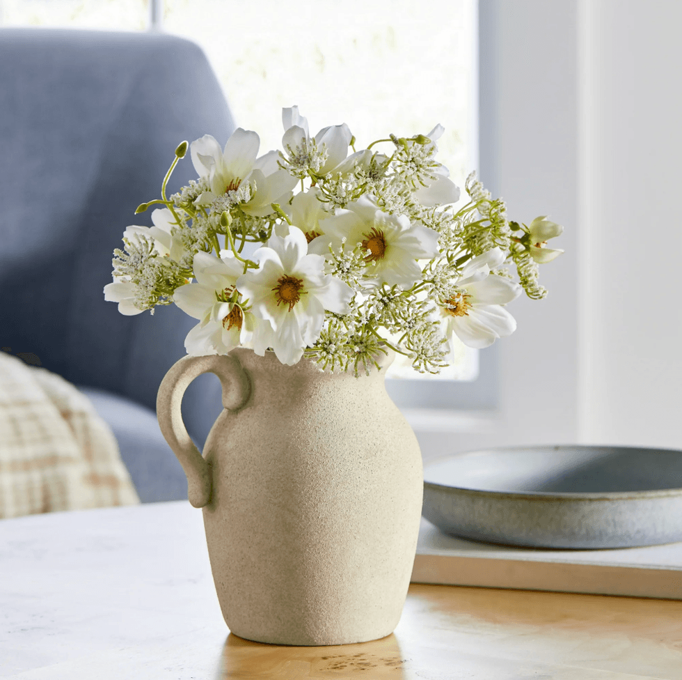 White flowers in a beige vase on a wooden table, with a blurred background.