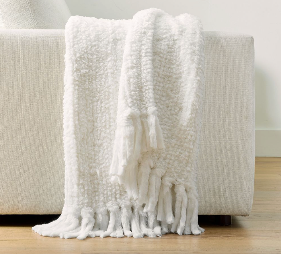 White fluffy throw draped over a light-colored couch, with tassels touching the wooden floor.