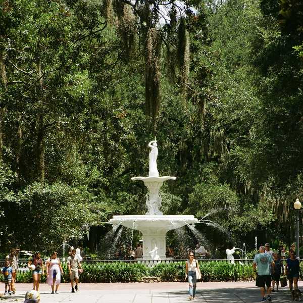 White fountain surrounded by trees and people in a lush park setting.