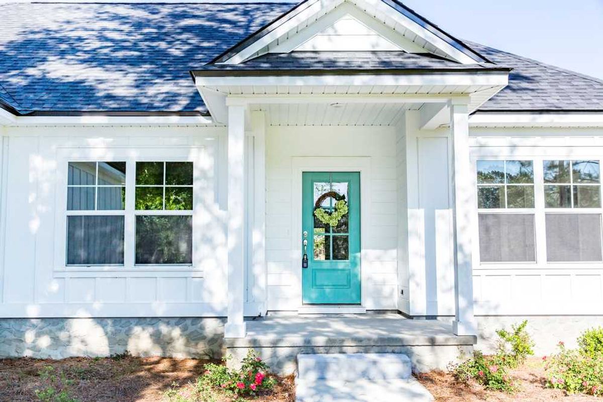 White house front with teal door, wreath, and large windows, surrounded by greenery.