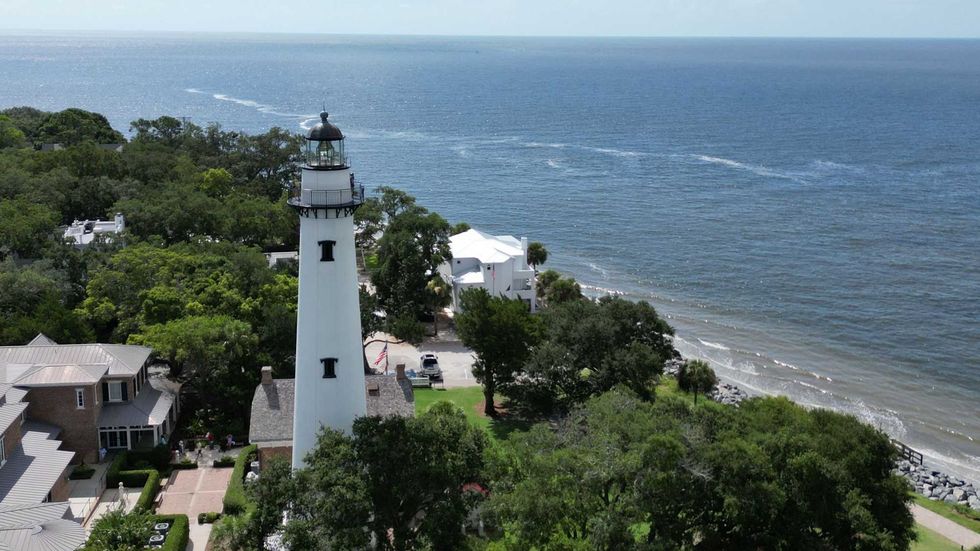 White lighthouse by the ocean, surrounded by trees and coastal buildings.