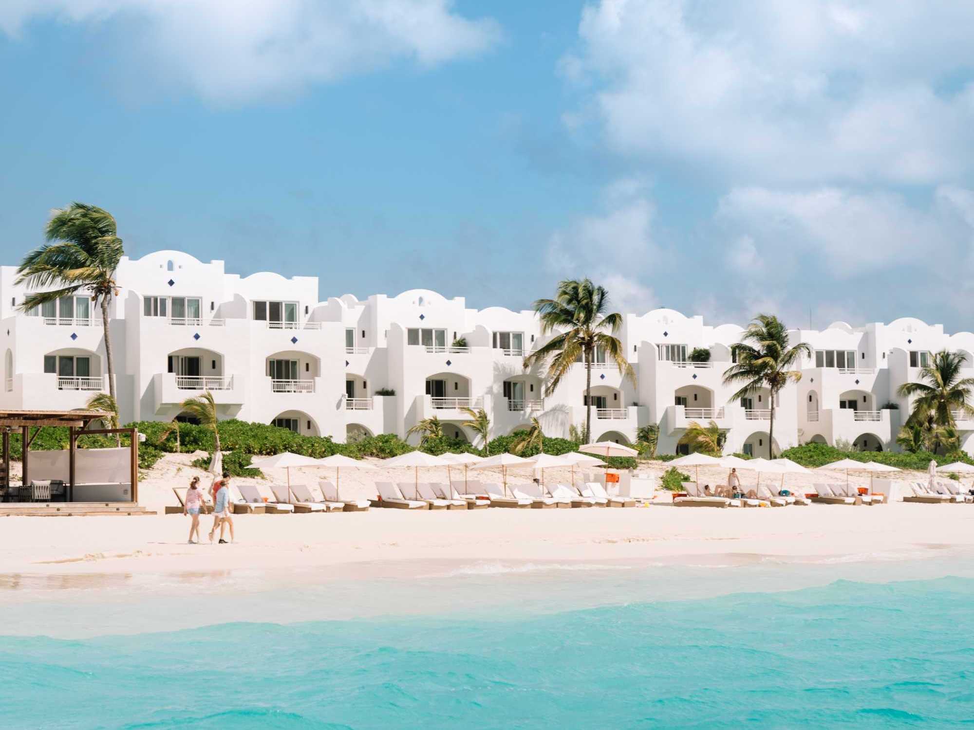 White resort by a sandy beach with palm trees, turquoise water, and blue sky.