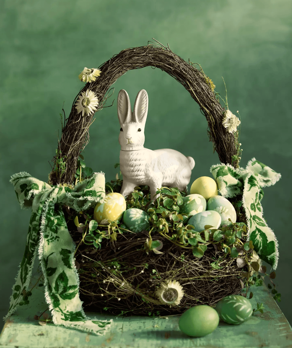 Wicker basket with a rabbit figurine, colored eggs, and greenery on a green background.