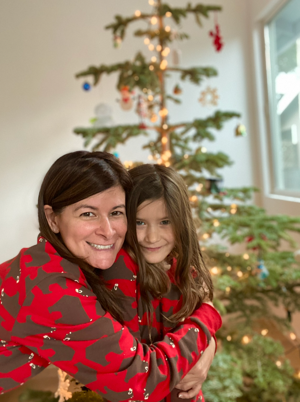 Woman and girl in matching pajamas hugging by a Christmas tree.