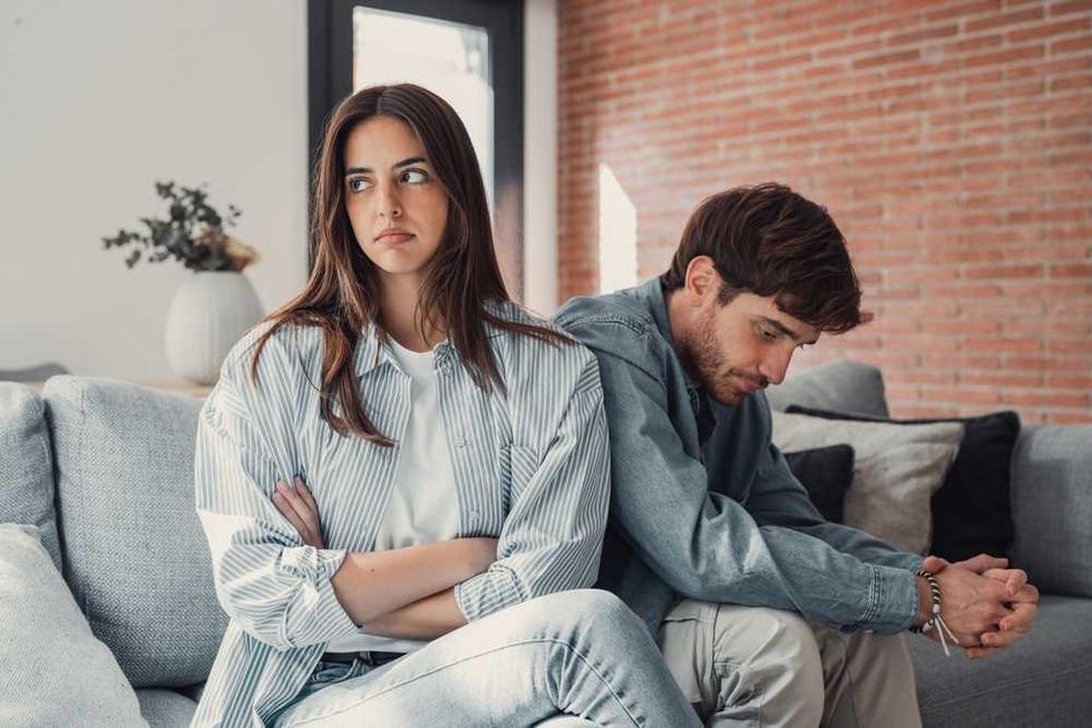 Woman and man sitting back-to-back on a couch, looking upset.