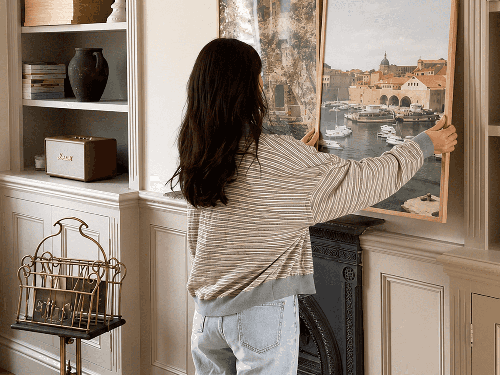 Woman arranging a framed harbor photo on a shelf near a vintage speaker.