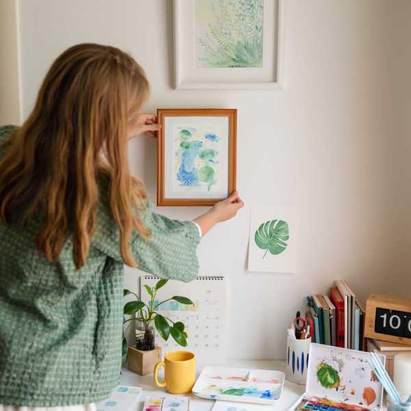 Woman arranging art on wall above a desk with art supplies and decor.