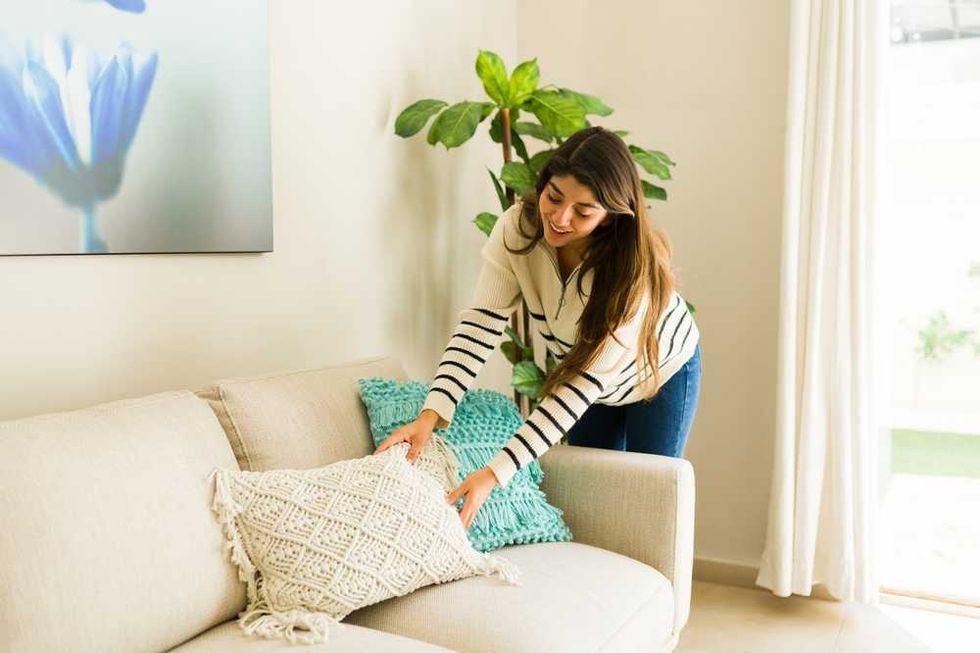 Woman arranging pillows on a beige sofa in a bright, cozy living room.