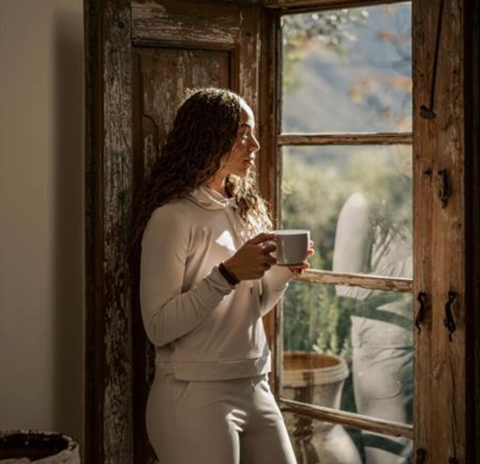 Woman drinking coffee and gazing out a rustic window in soft natural light.