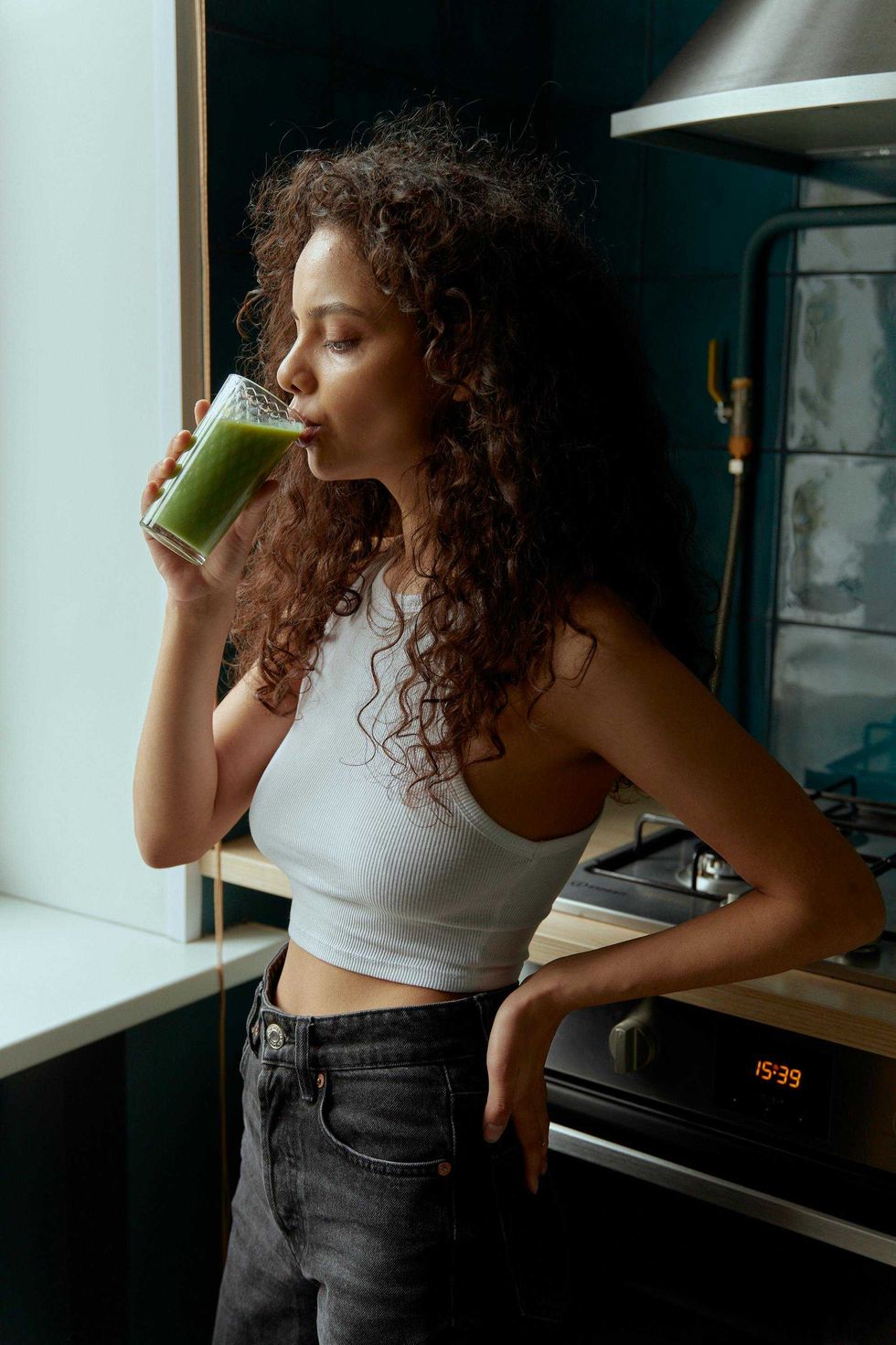 Woman drinking green juice in a kitchen, wearing a white top and black jeans.