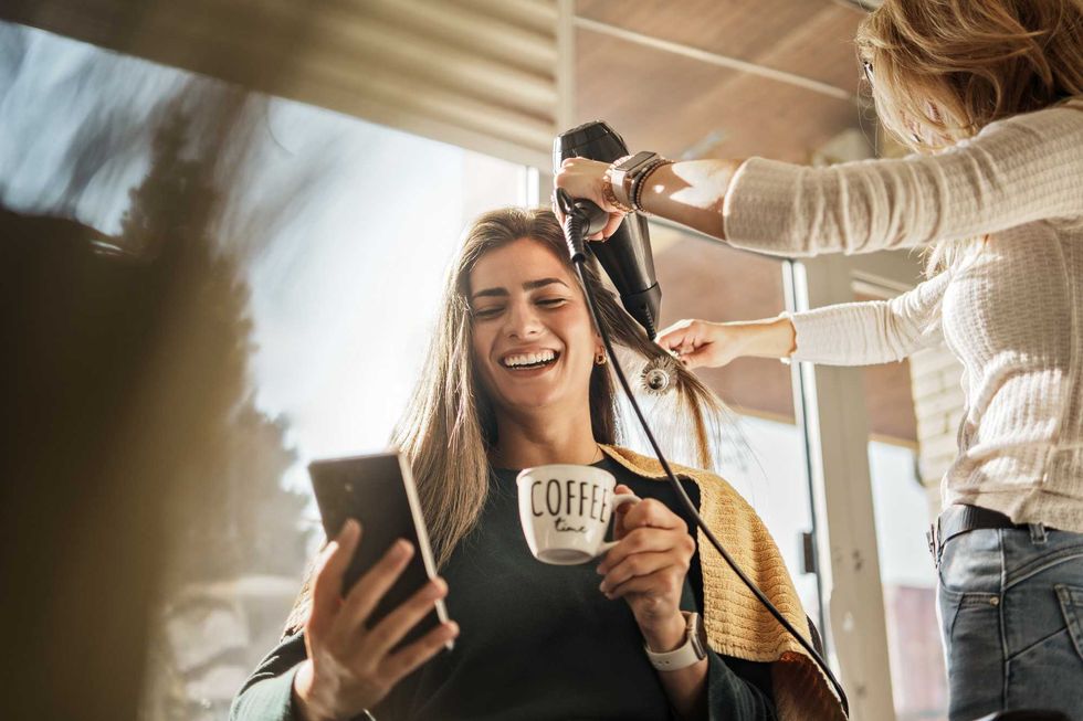 Woman enjoys coffee and phone at salon while getting hair blow-dried.