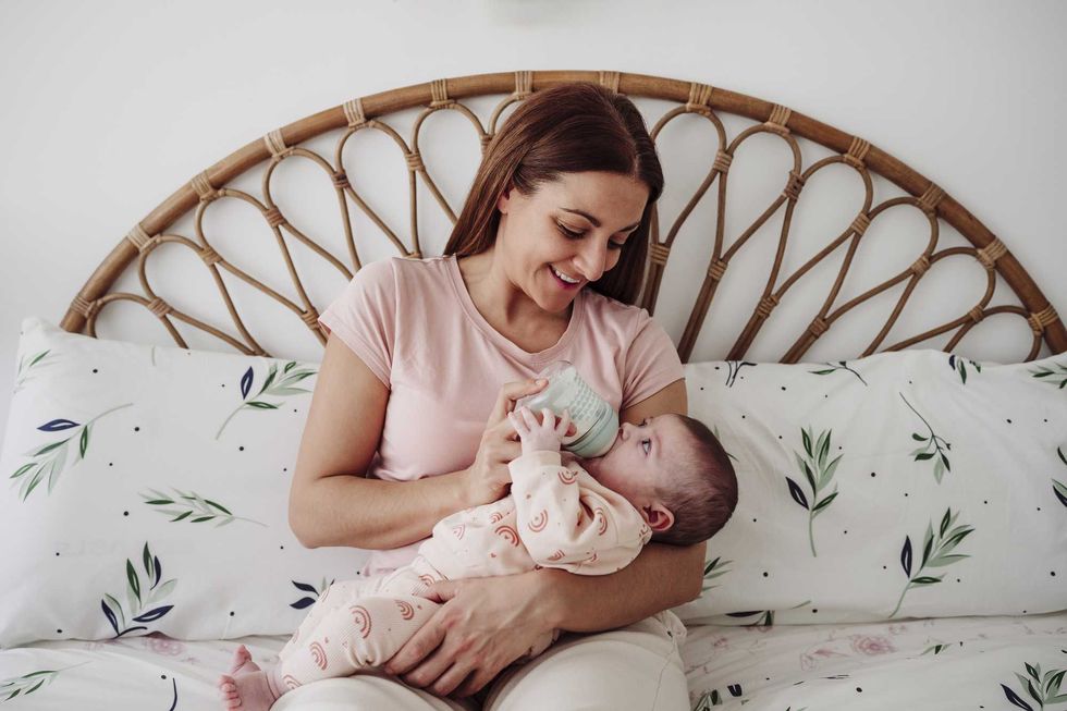 Woman feeding baby with bottle on a floral-patterned bed.