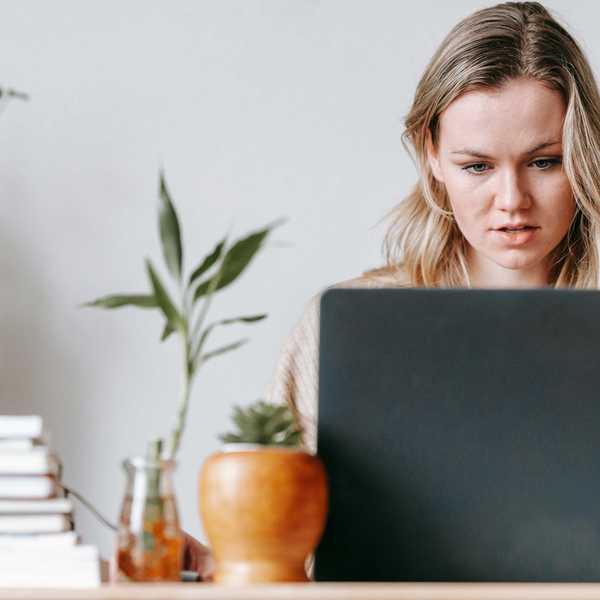 Woman focused on laptop at desk with plants and books.