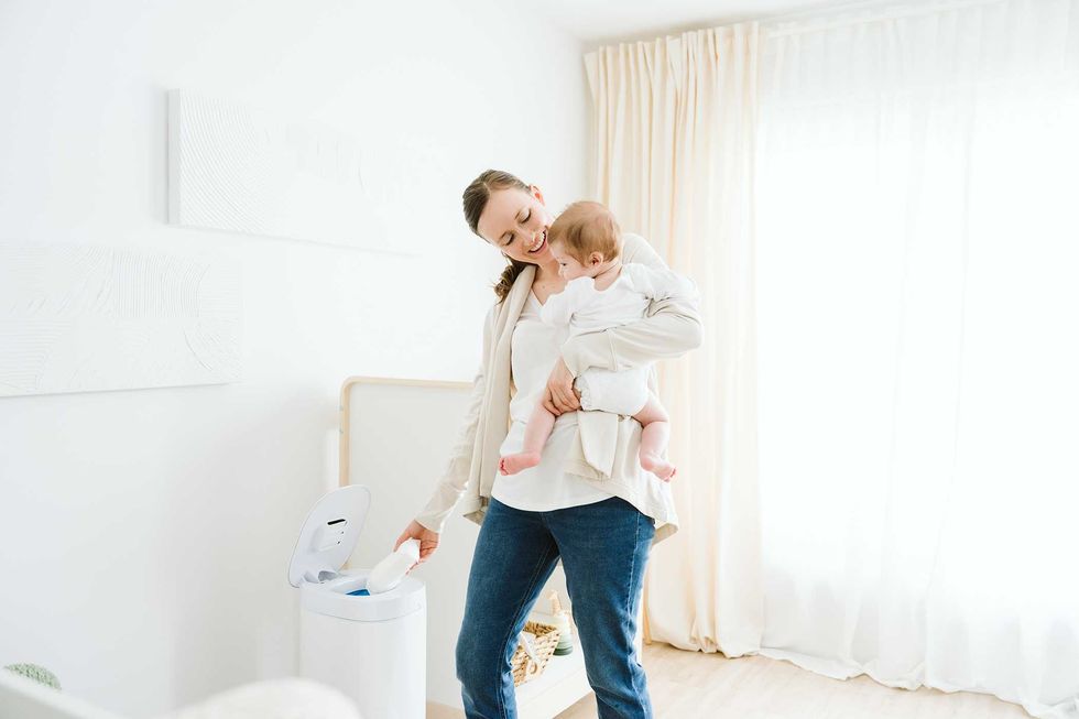 Woman holding baby, smiling, while disposing diaper in a white nursery.