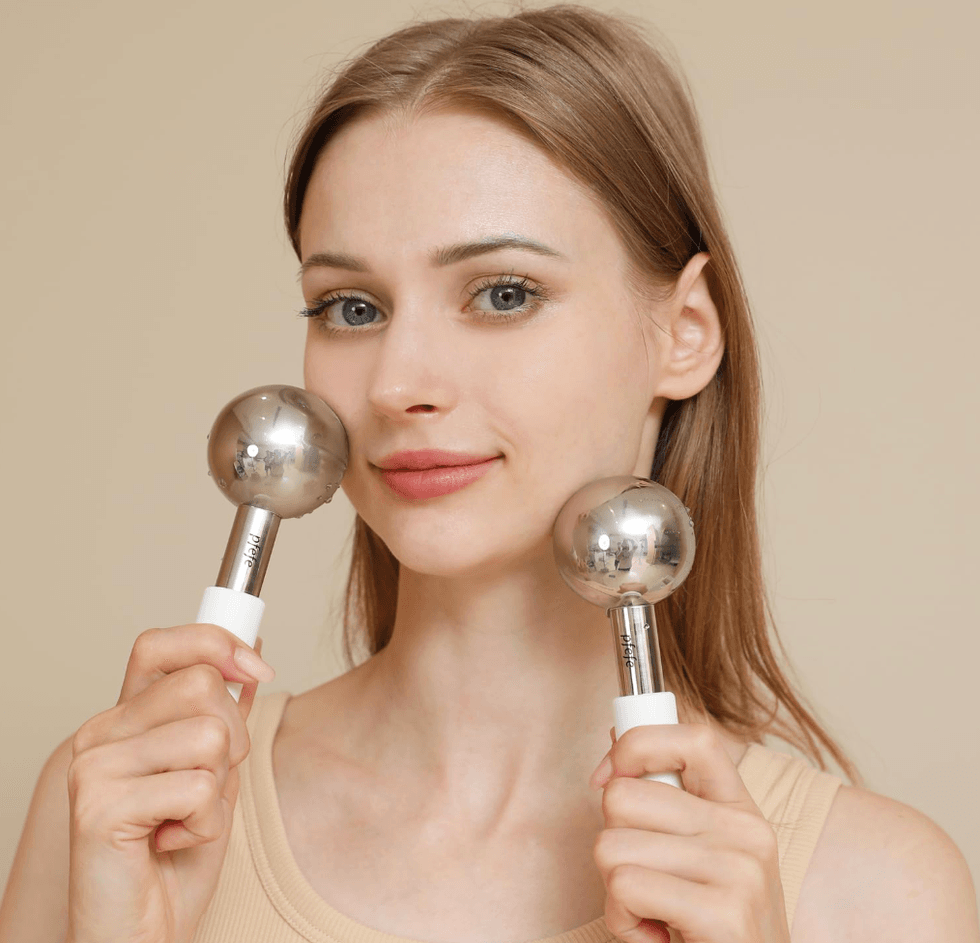 Woman holding two silver facial massage tools near her cheeks.