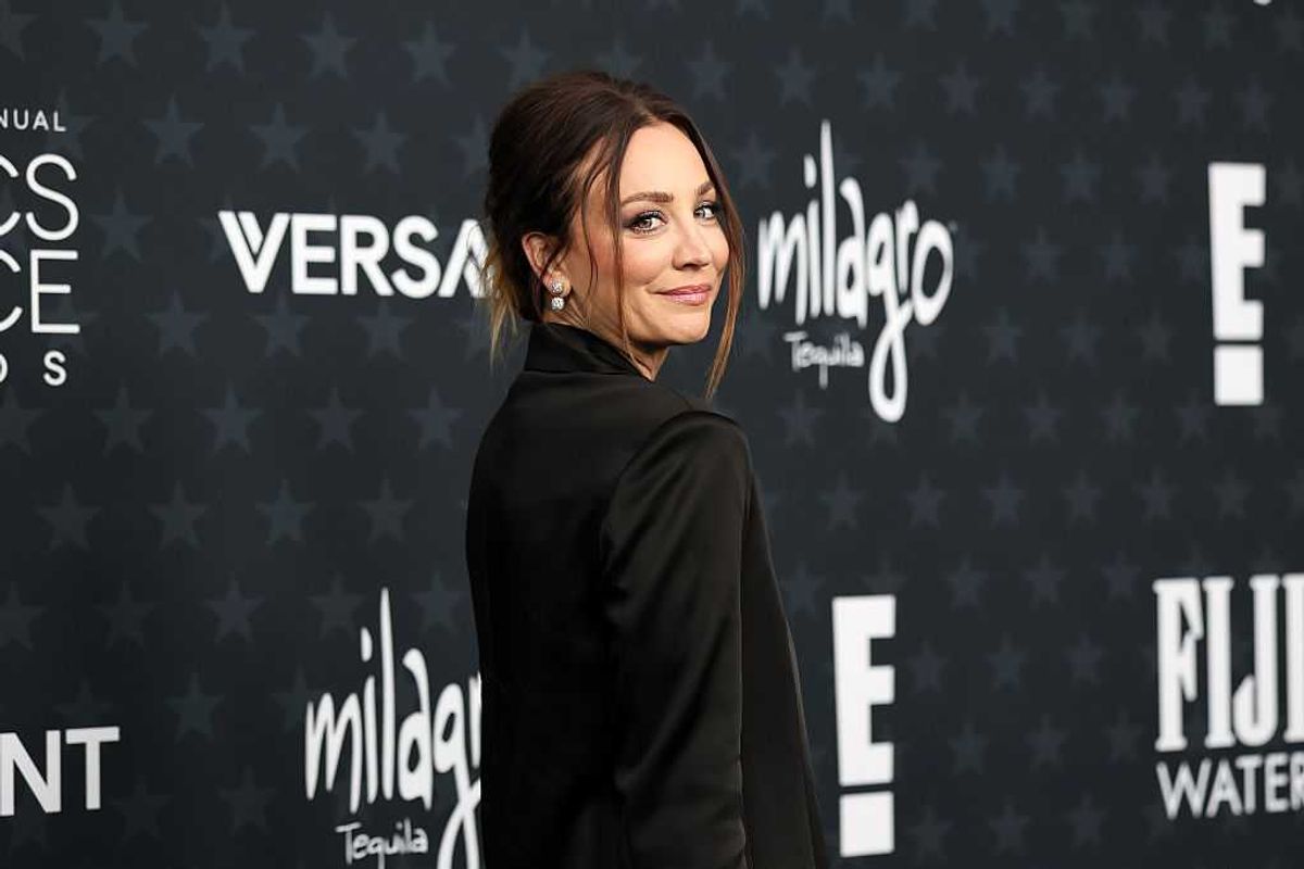 Woman in a black outfit poses at an event with sponsor logos on a dark background.