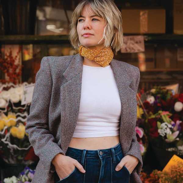 Woman in a blazer and scarf outside a flower shop, hands in pockets, looking to the side.