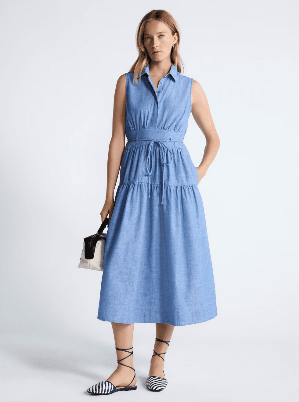 Woman in a blue sleeveless dress holding a handbag, wearing striped flats.
