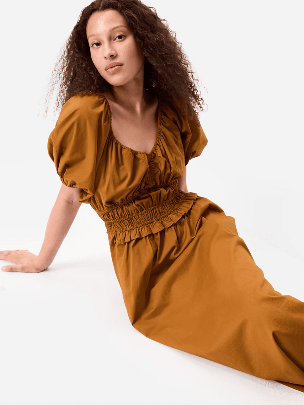 Woman in a brown dress sitting, against a white background.