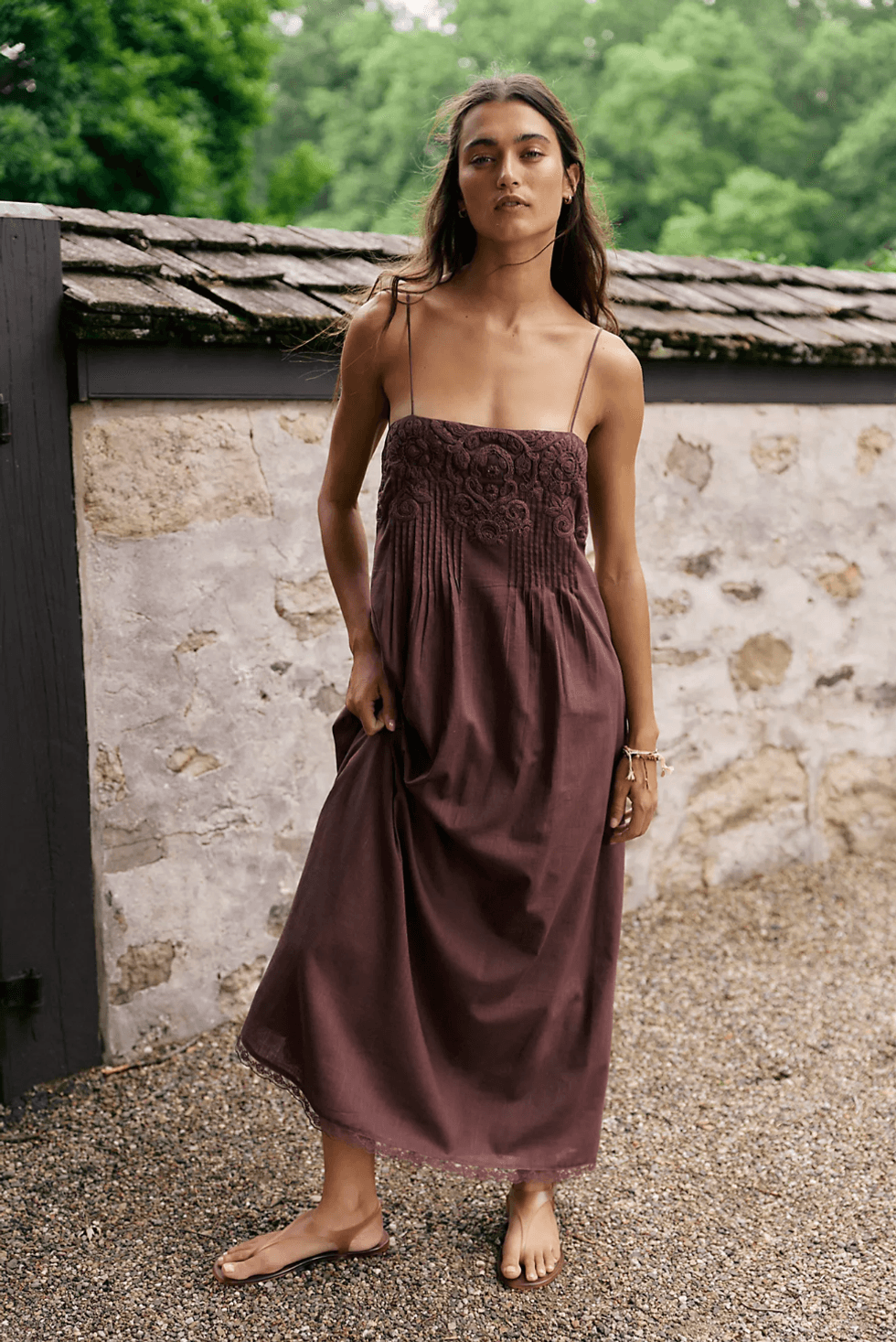 Woman in a brown dress standing outdoors near a stone wall.