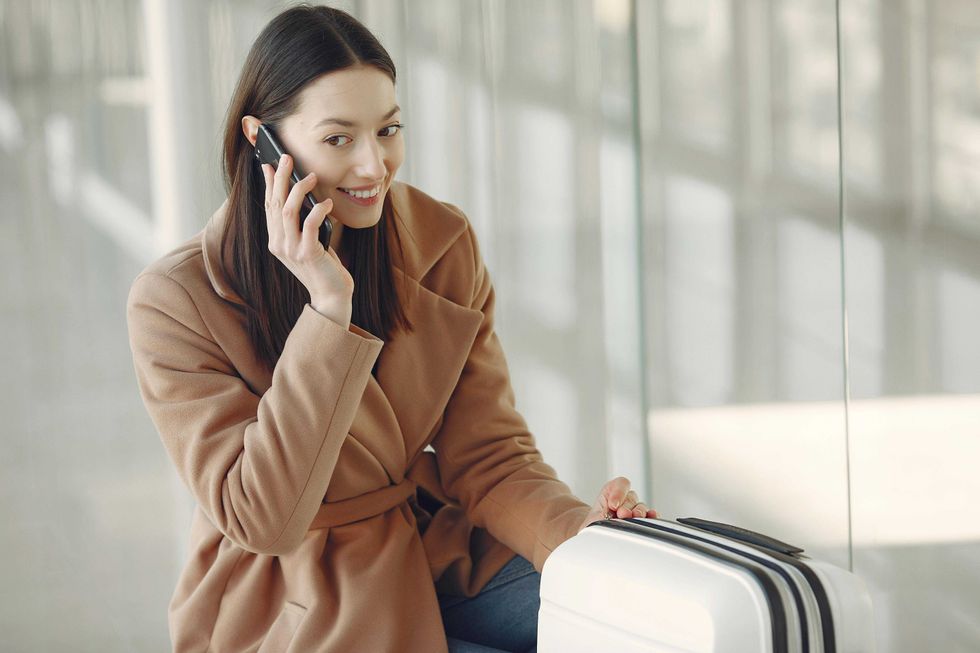 Woman in a coat sits by suitcase, talking on phone in a bright glass-walled area.