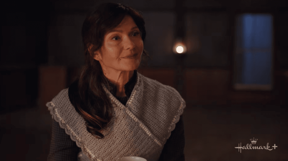 Woman in a cozy sweater holds a mug, standing indoors by soft, warm lighting.