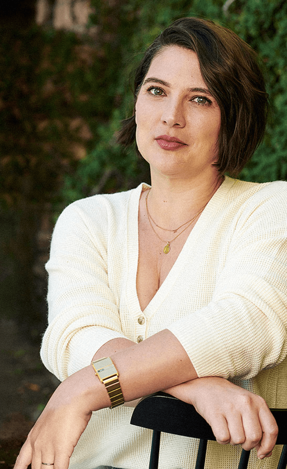 Woman in a cream sweater sitting outside with greenery in the background.