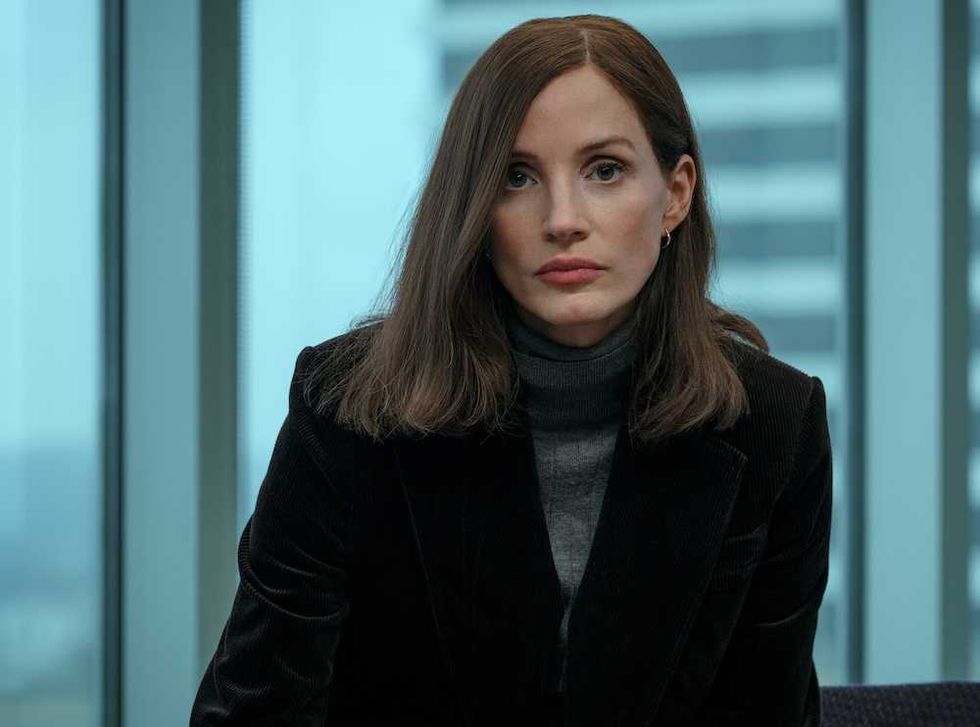 Woman in a dark blazer and turtleneck, sitting indoors, looking forward with a neutral expression.