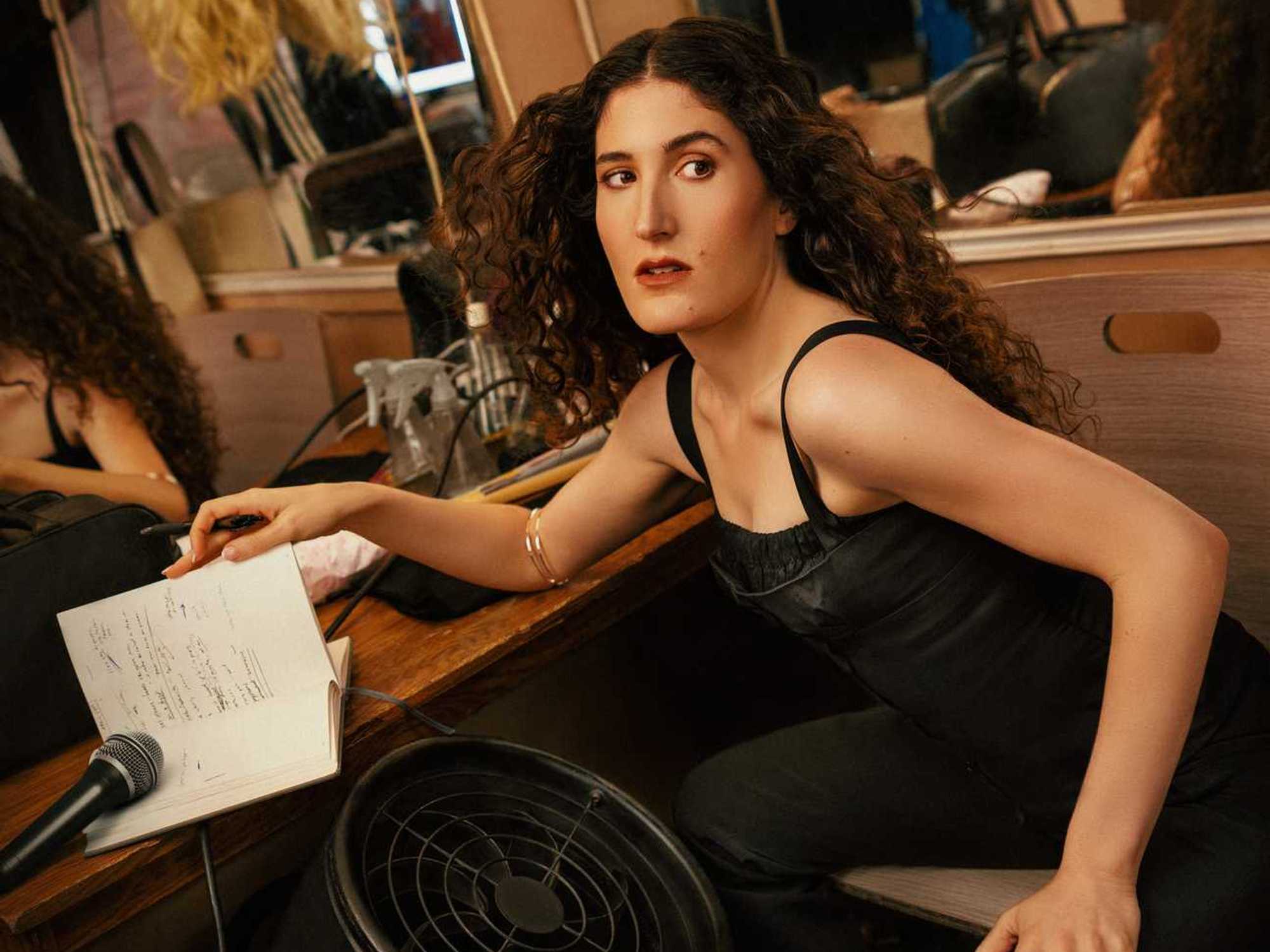 Woman in a dressing room with notebook and microphone, looking over shoulder.