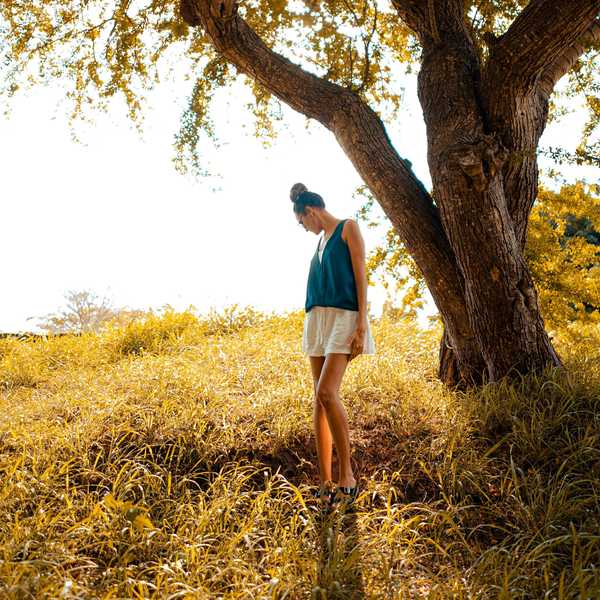 Woman in a field under a large tree, wearing a blue top and white shorts, looks down.