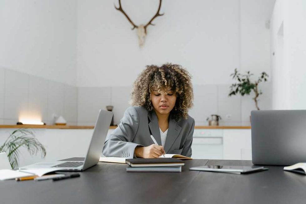 Woman in a gray suit writing at a desk with laptops in a modern office setting.