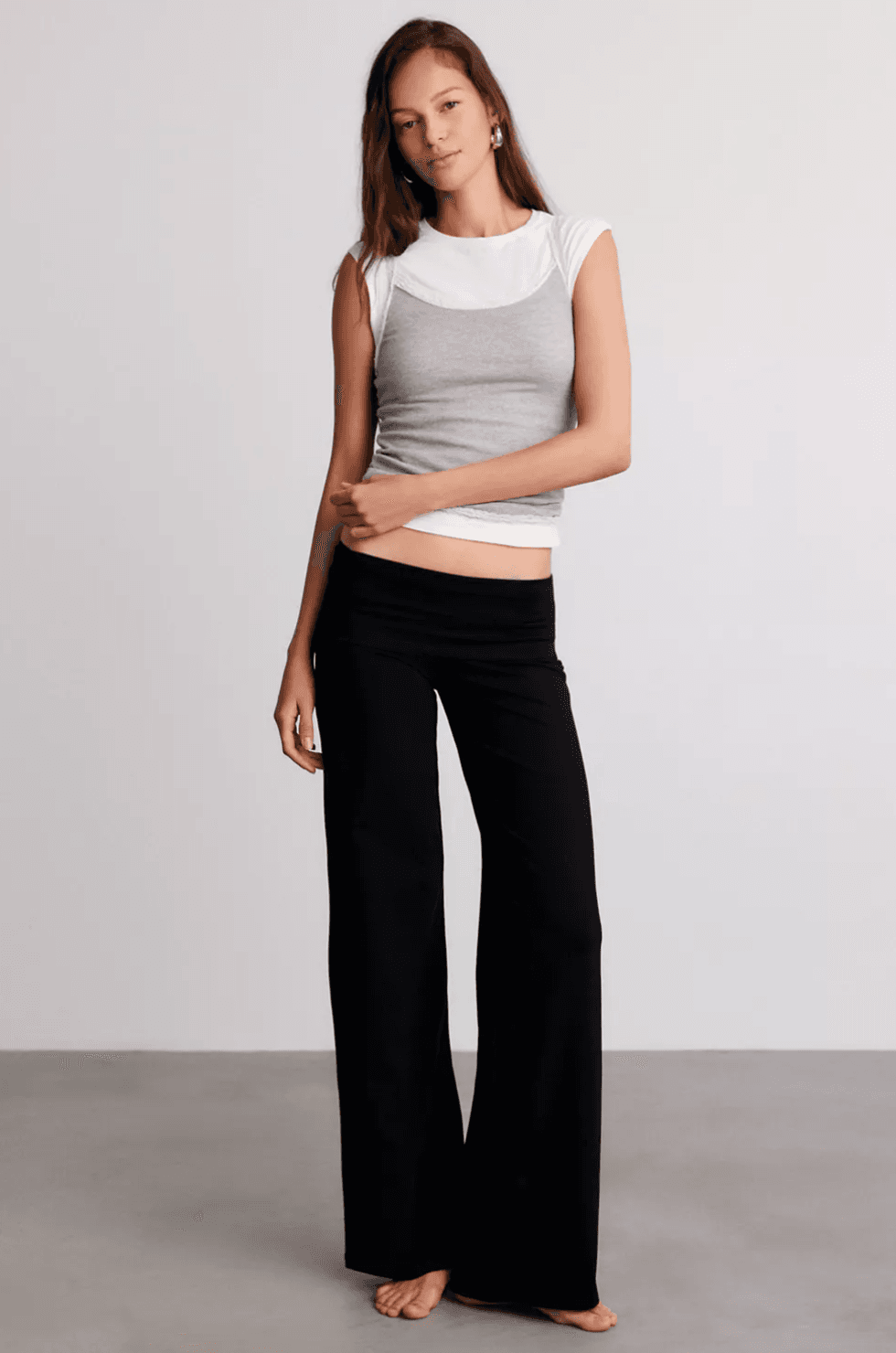Woman in a gray tank top and black pants, standing barefoot on a gray floor.