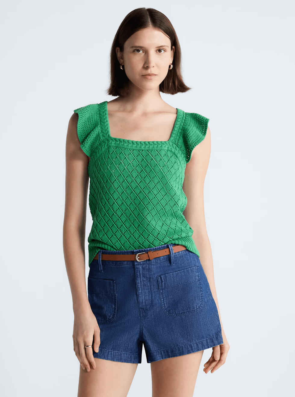 Woman in a green lace top and blue shorts with a brown belt, standing against a plain background.