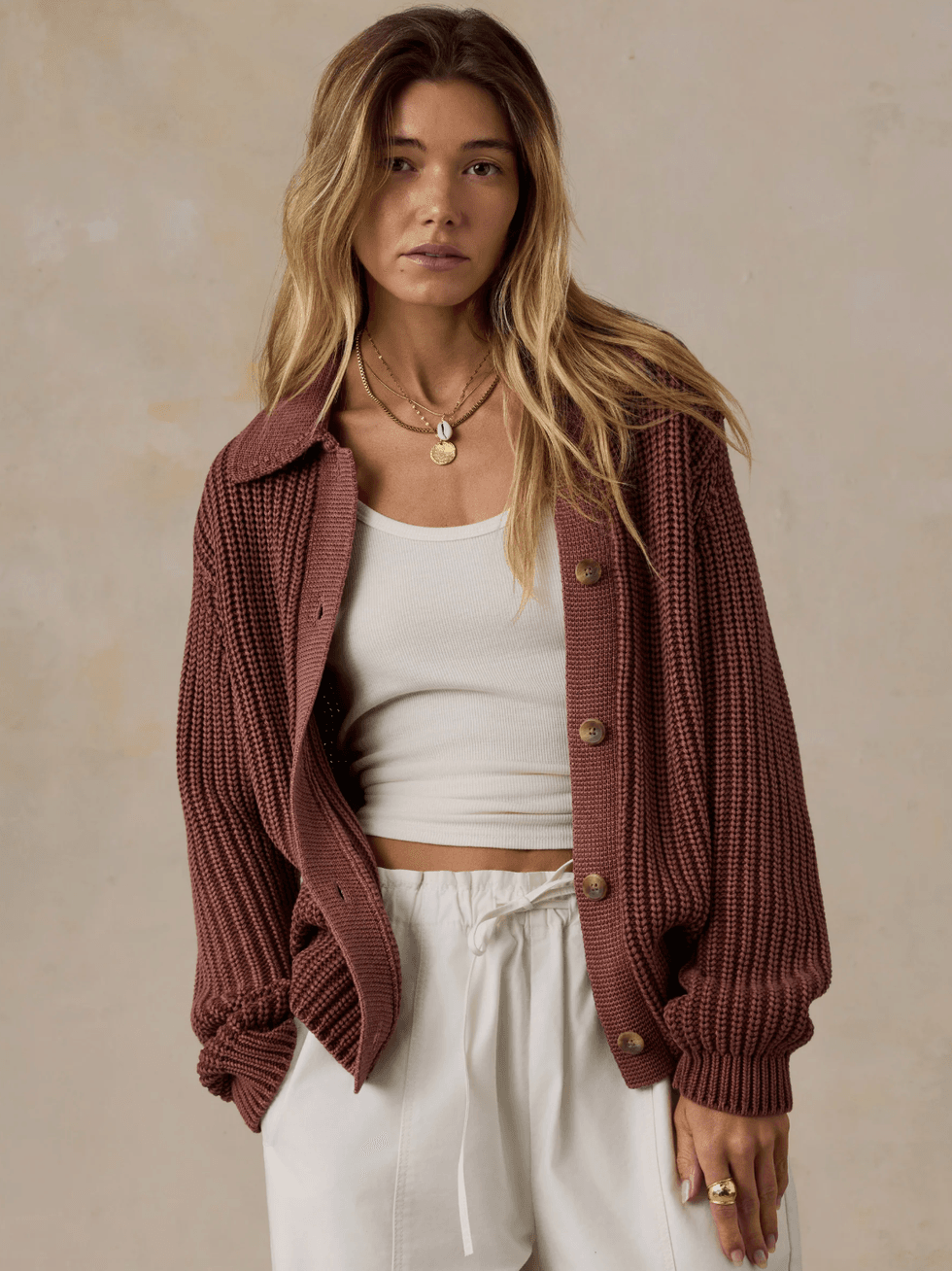 Woman in a maroon cardigan over a white tank top, wearing white pants.