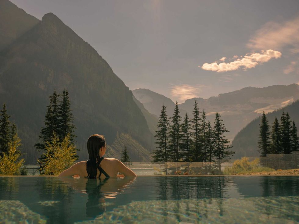 Woman in a pool, overlooking a mountain lake and forest under a cloudy sky.